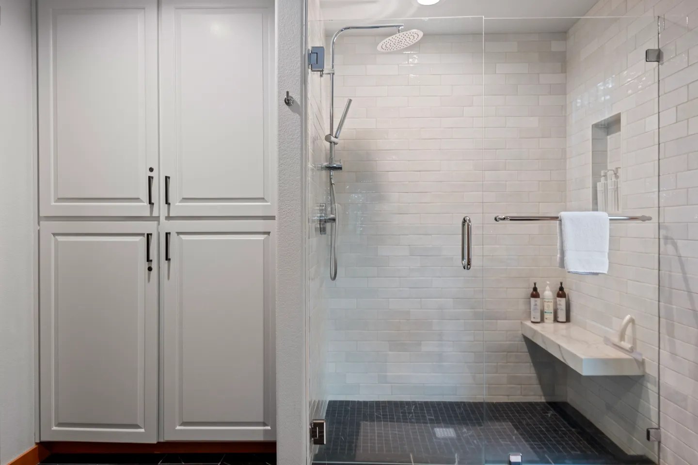 Walk-in shower with white subway tile, glass door, bench seat, and adjacent white storage cabinet.
