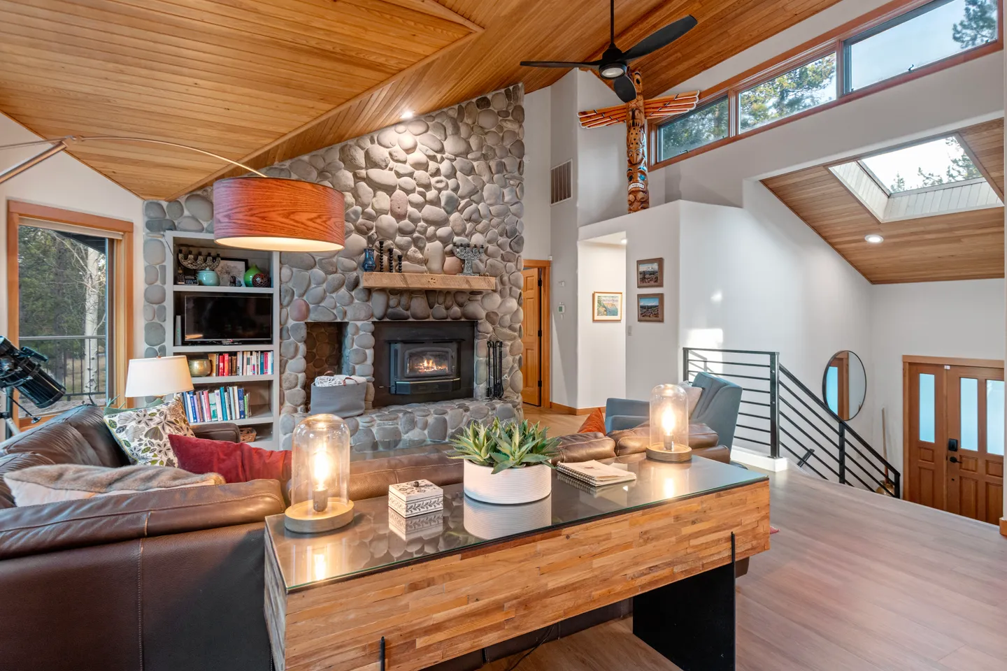 Living room with wood-paneled angled ceilings, a stone fireplace, leather sectional sofa, and view towards the entryway stairs.