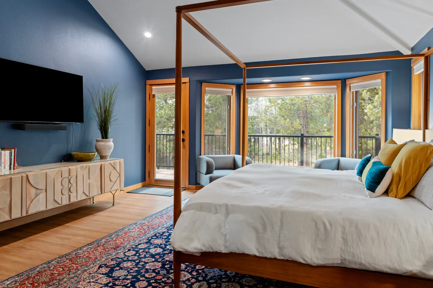 Bedroom featuring a wooden four-poster canopy bed, blue walls, a flat-screen television mounted above a light wood console, and a door leading to a balcony.