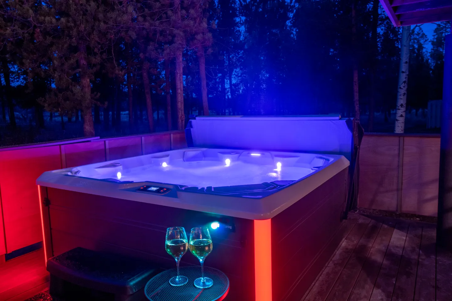 An outdoor hot tub illuminated at night with colored lights on a wooden deck surrounded by trees.