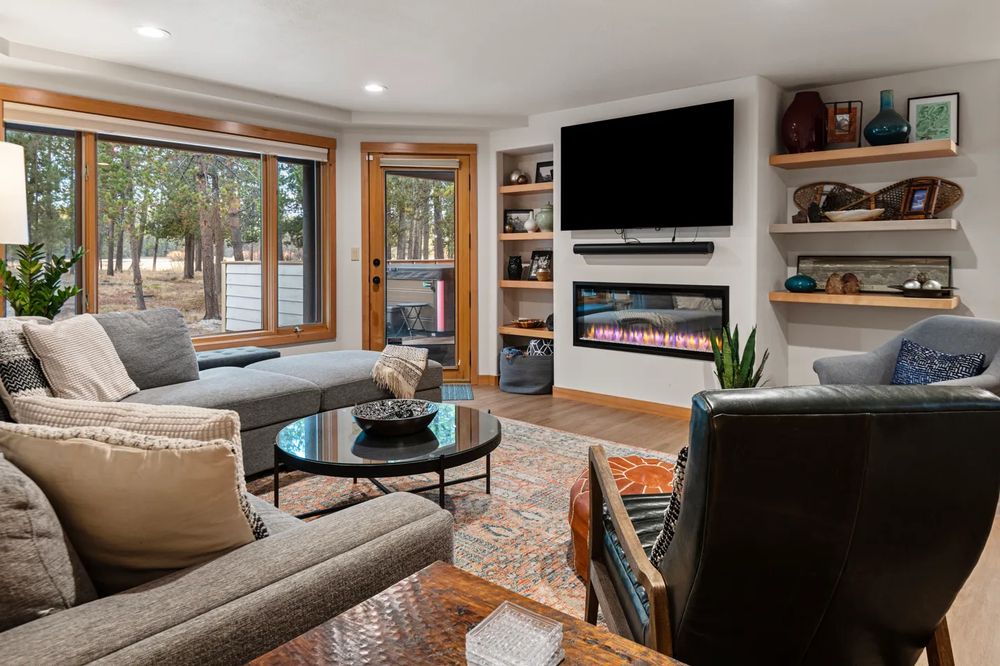 Living room with a sectional sofa, armchairs, modern electric fireplace, wall-mounted TV, and large windows looking out to trees.
