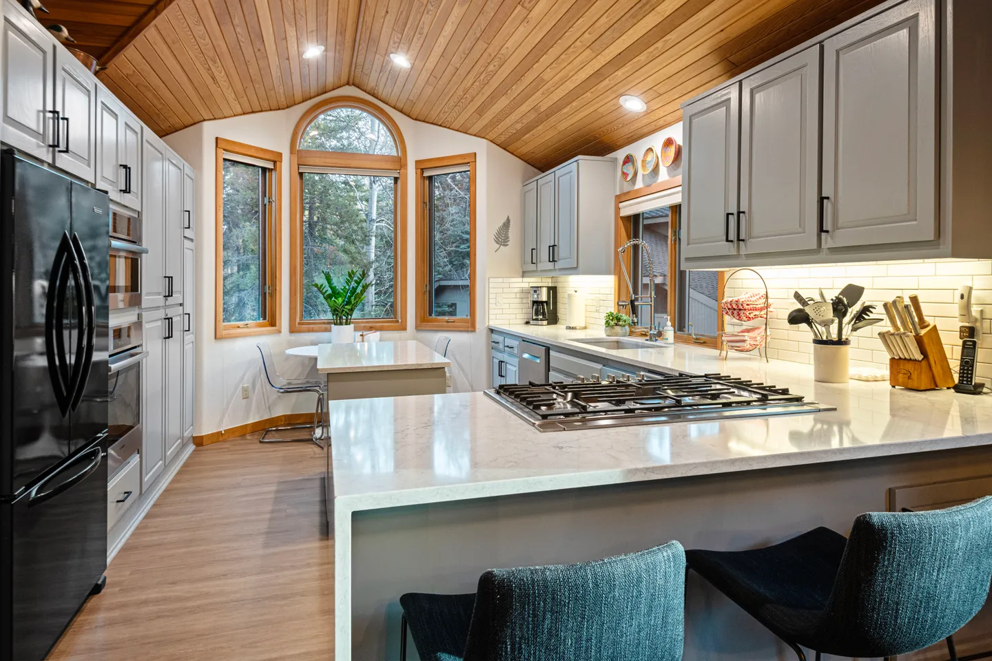 A kitchen with light gray cabinetry, light countertops, wood-paneled vaulted ceiling, and a breakfast nook with an arched window.