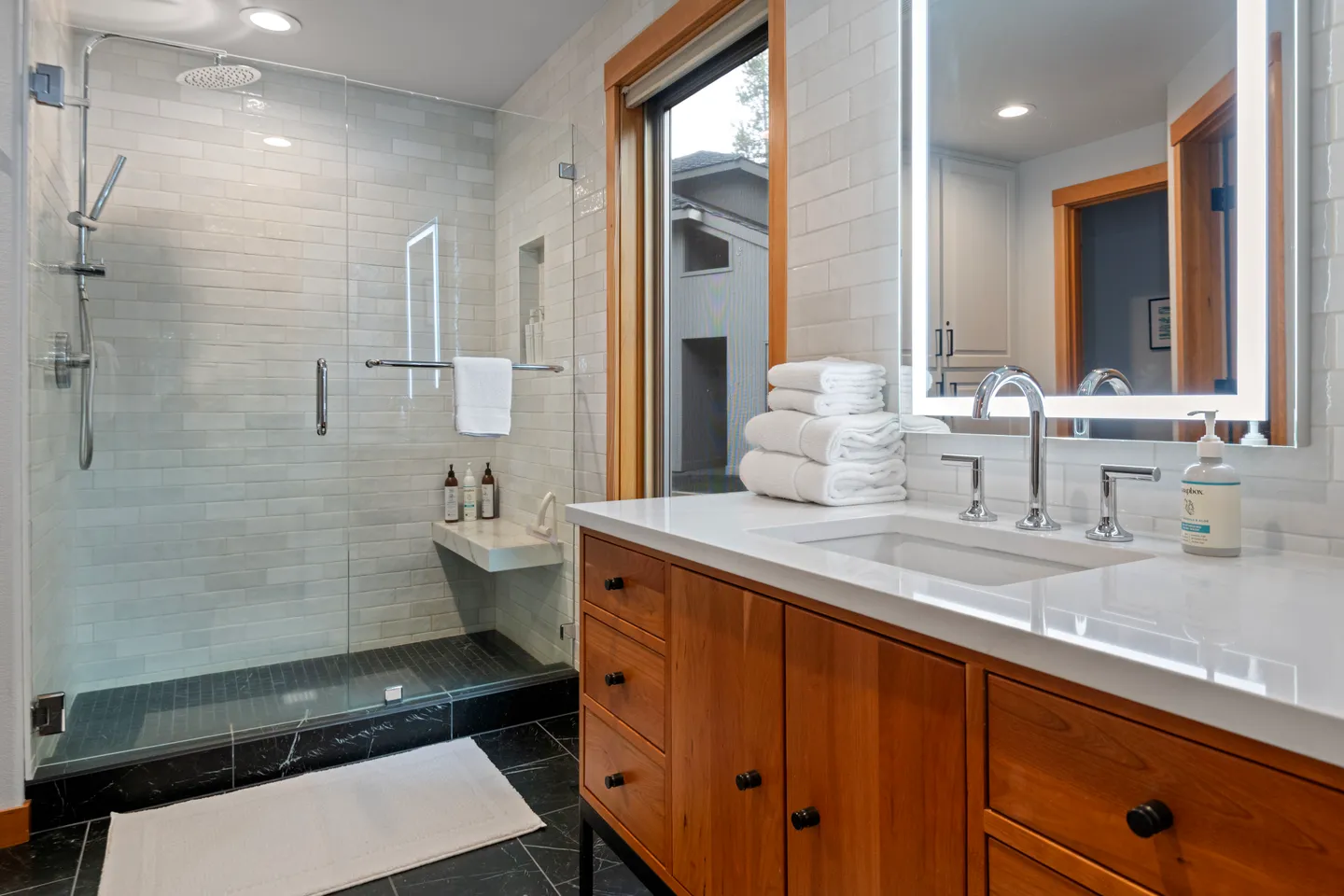 Bathroom with a glass-enclosed walk-in shower and a wooden vanity with a white countertop and backlit mirror.