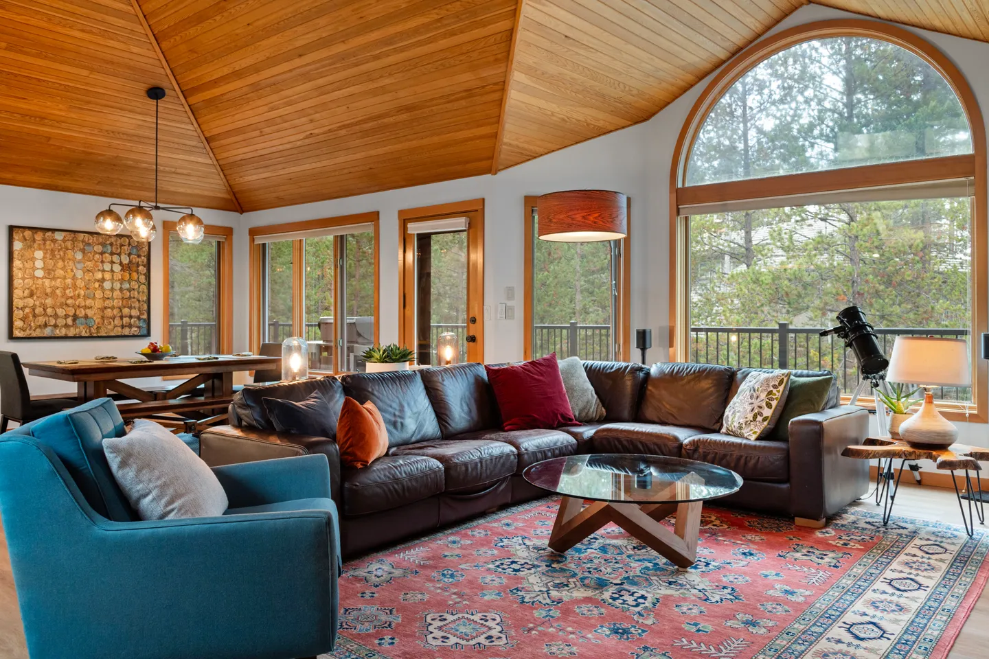 Living room with vaulted wood-paneled ceiling, large arched window, leather sectional sofa, patterned area rug, and dining area in the background.