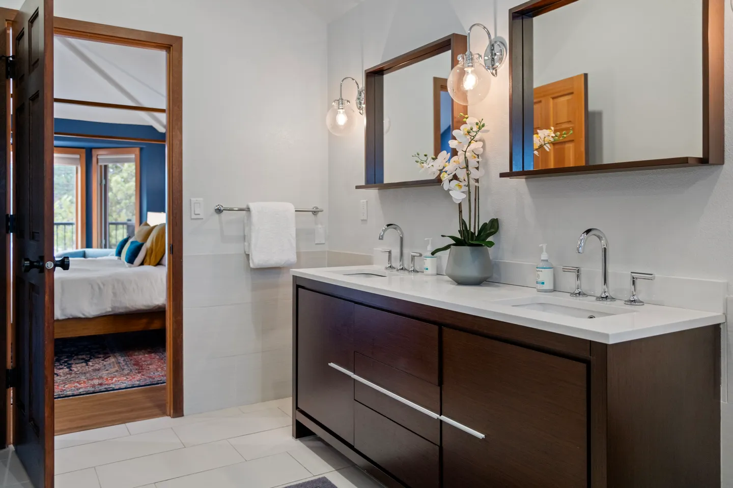 Bathroom with a double-sink dark wood vanity, white countertops, two framed mirrors with sconces, and an open doorway leading to a bedroom.