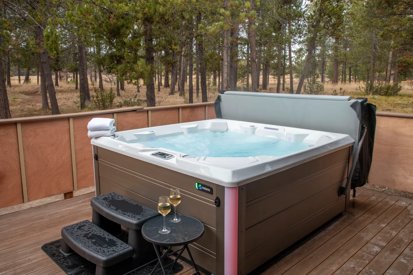 Outdoor hot tub on a wooden deck overlooking a pine forest with two wine glasses on a side table.
