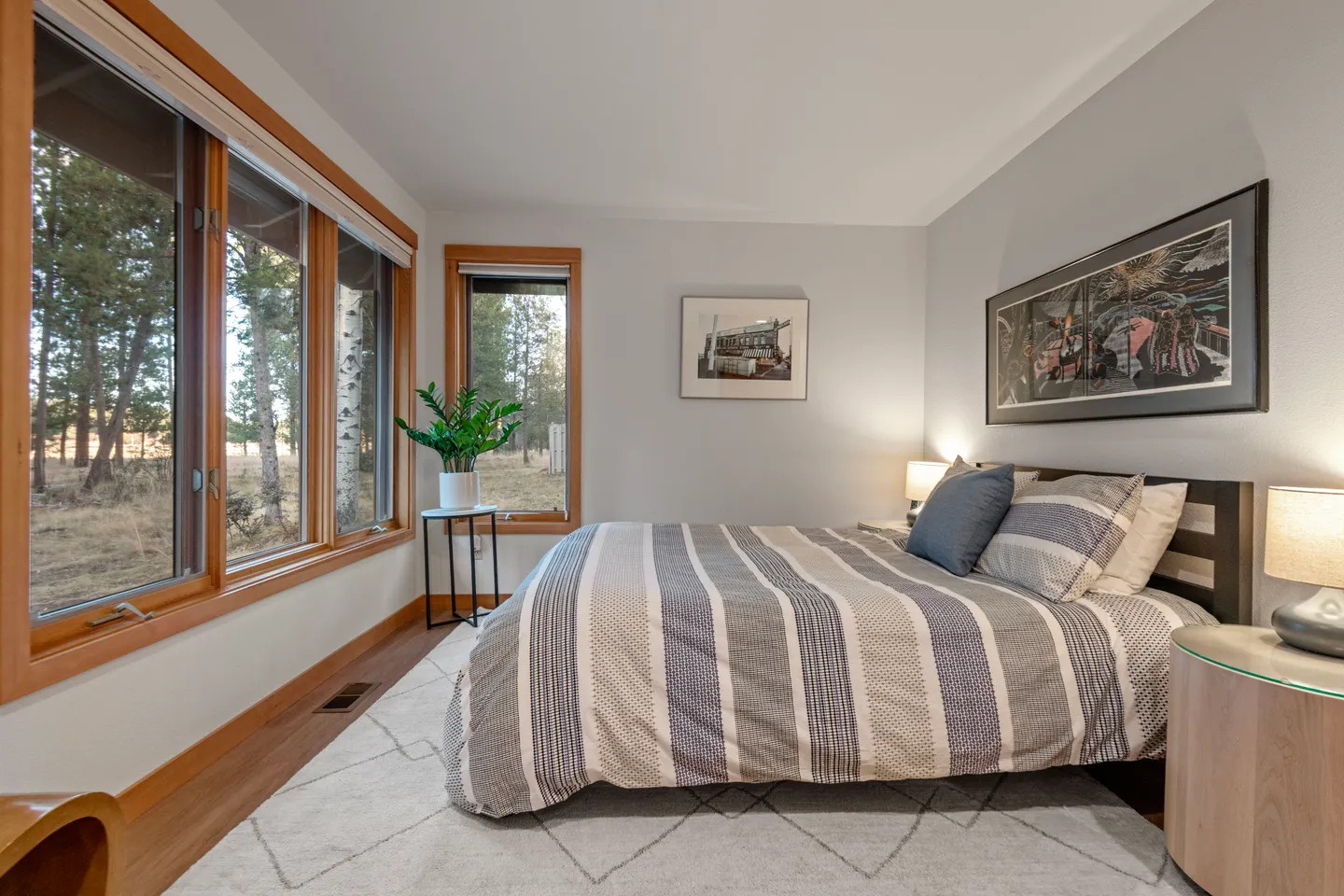 Bedroom with a striped bedspread, light-colored walls, nightstands with glowing lamps, and large wood-framed windows looking out to trees.