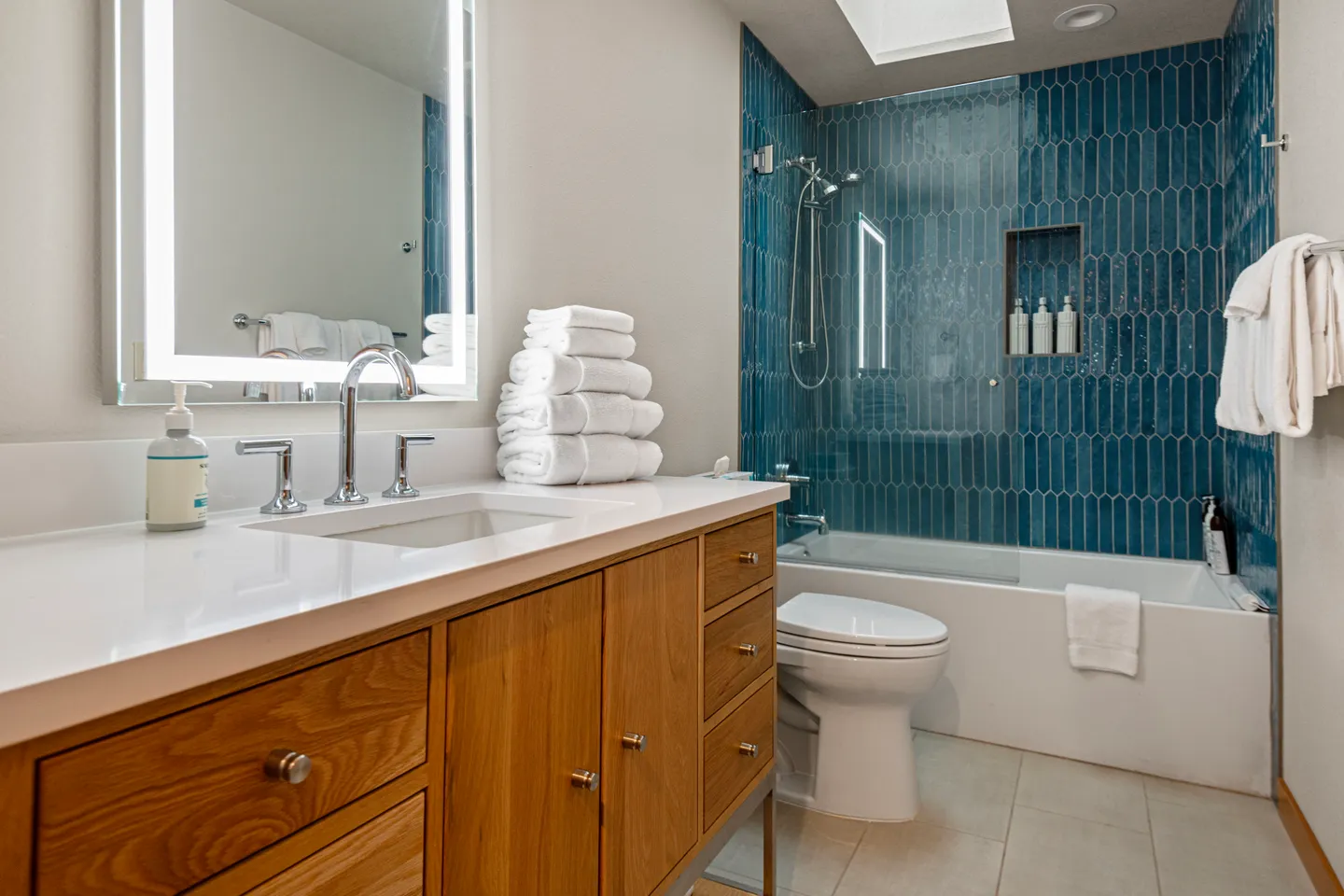 Bathroom with a wood vanity, white countertop, illuminated mirror, toilet, and combination bathtub and shower lined with blue picket tiles.