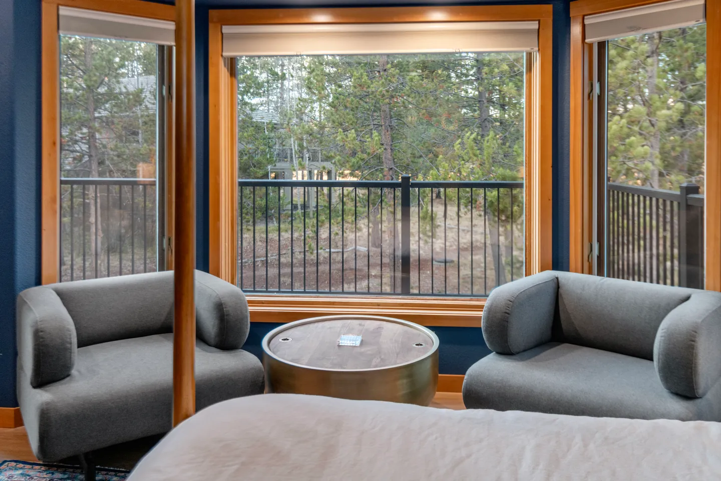 Seating area in a bedroom featuring two gray armchairs, a round coffee table, and large bay windows overlooking pine trees.
