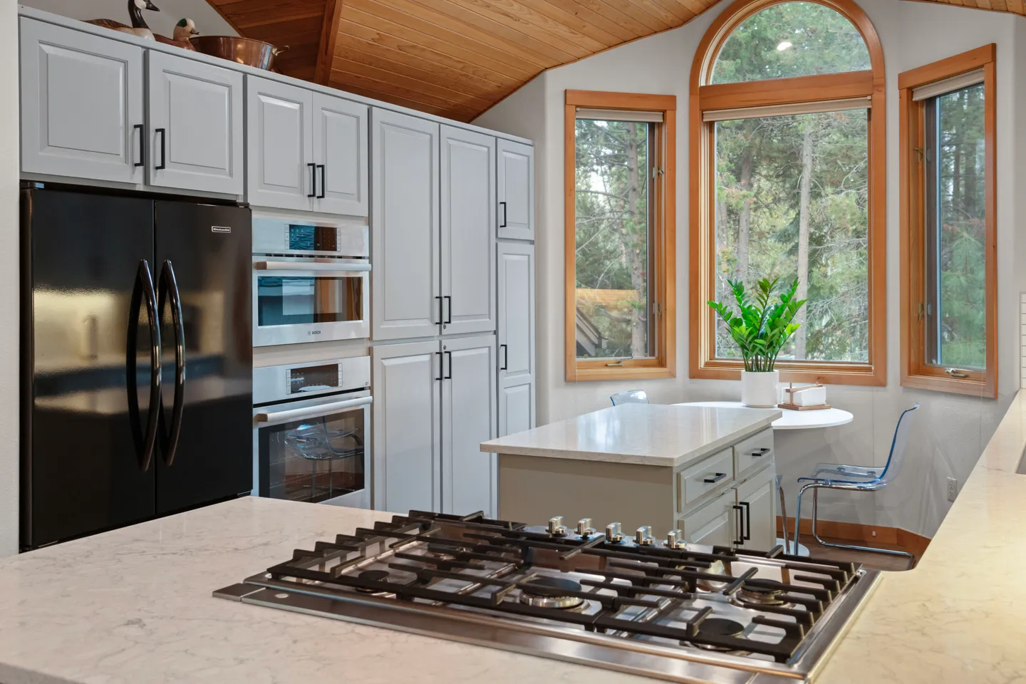 Kitchen featuring light gray cabinetry, built-in double ovens, a gas cooktop on the island, and a casual dining table by arched wood-framed windows.