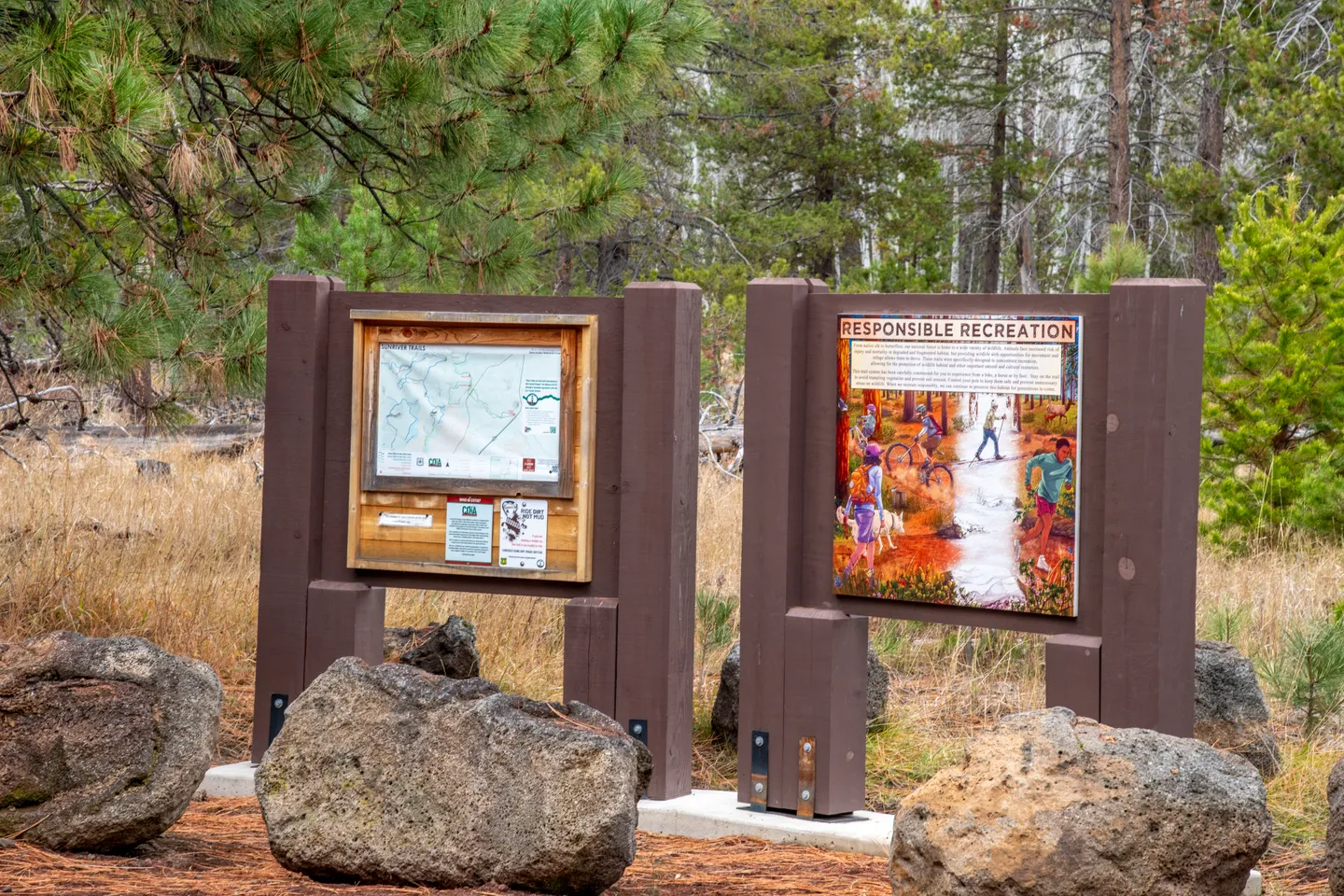 Two outdoor informational trail signs mounted on wooden posts surrounded by boulders and pine trees.