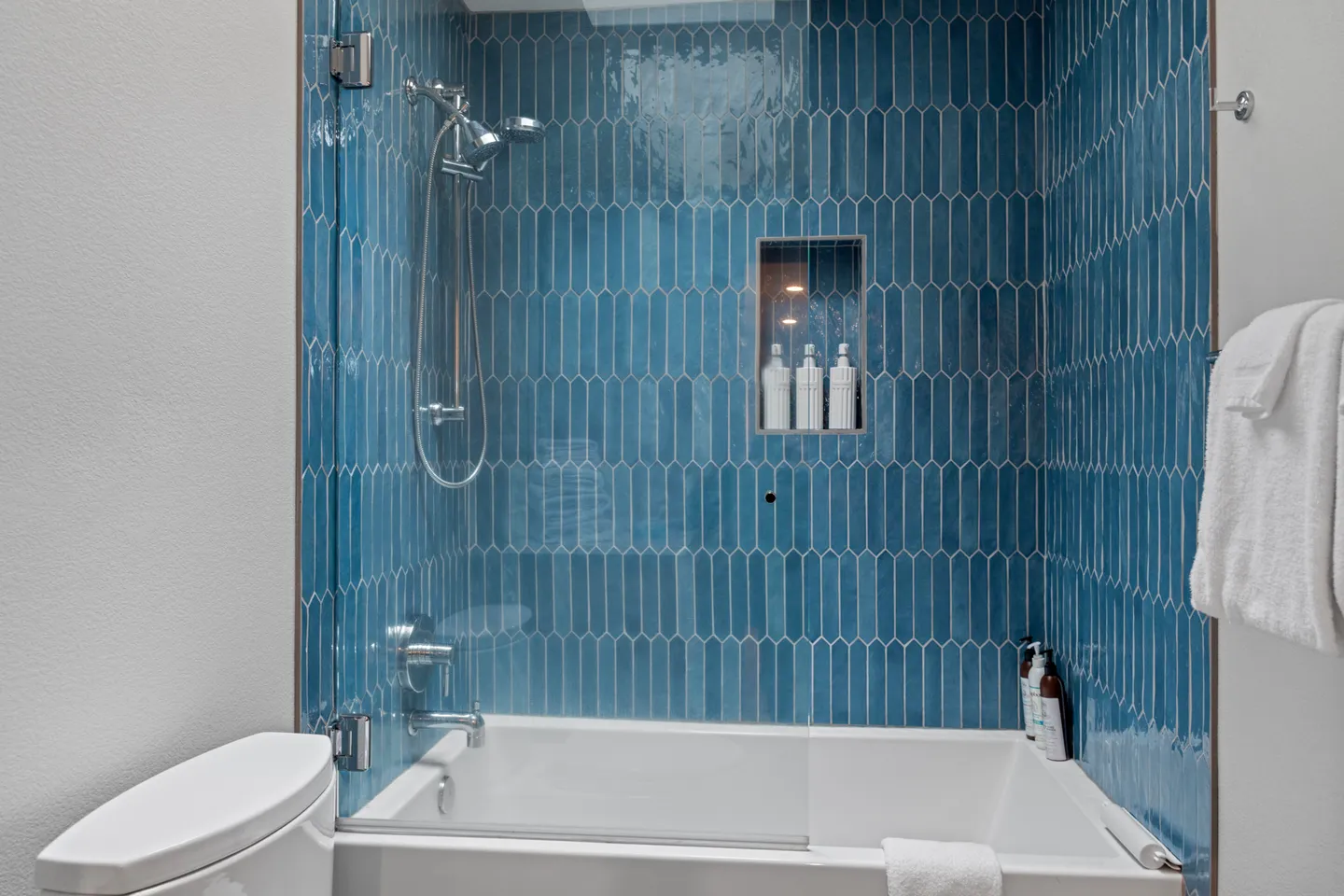 A bathtub and shower combination featuring glossy vertical blue subway tile and a glass partition.