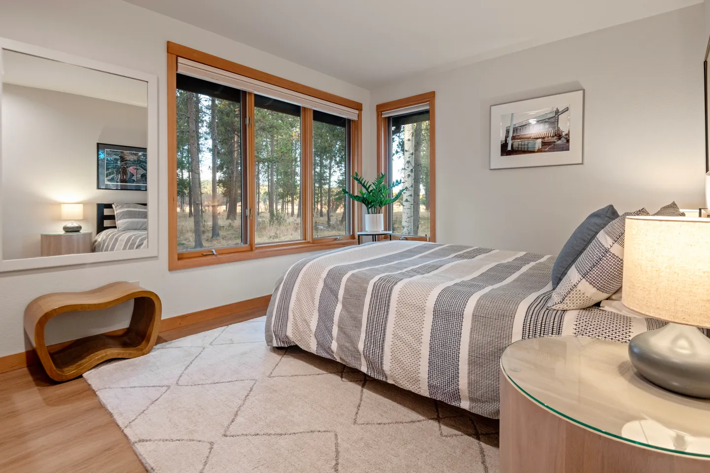Bedroom with a striped comforter, large wood-framed windows looking out to trees, an area rug, and a wall mirror.