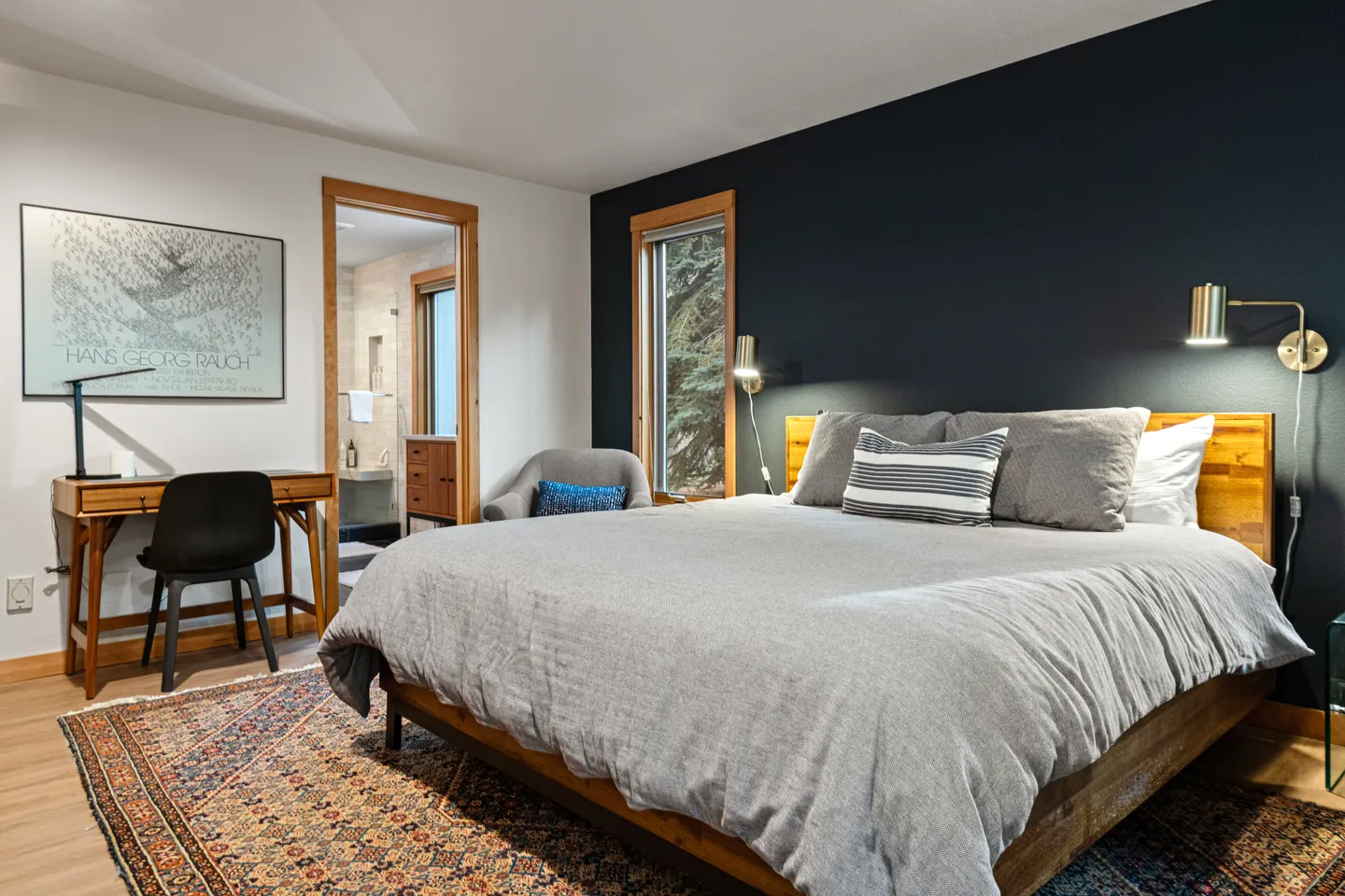 Bedroom featuring a made wooden bed against a dark accent wall, flanked by reading sconces, with a small desk and a view into the en-suite bathroom.