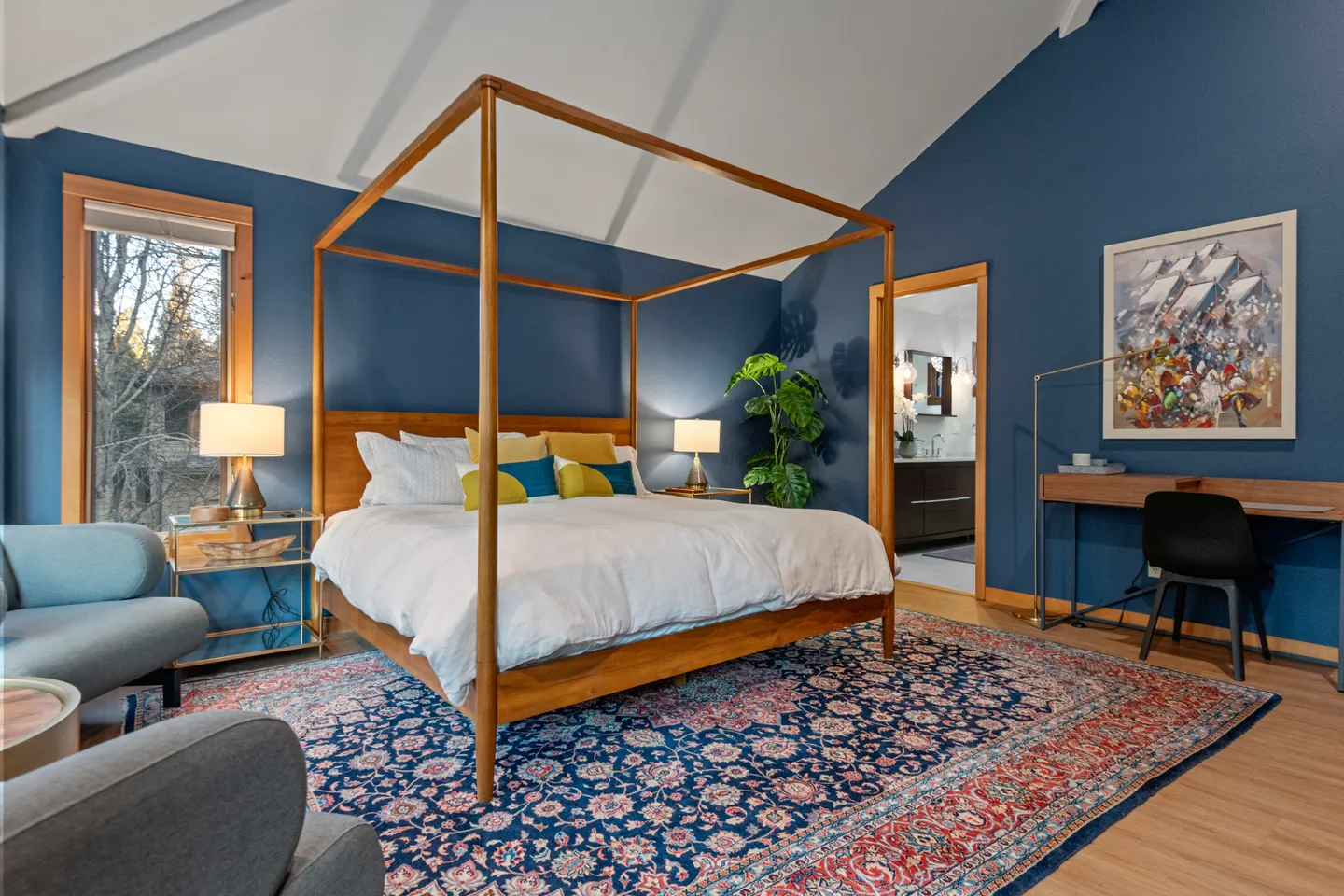 Bedroom with deep blue walls, a wooden canopy bed, vaulted ceiling, and an en-suite bathroom visible through an open doorway.