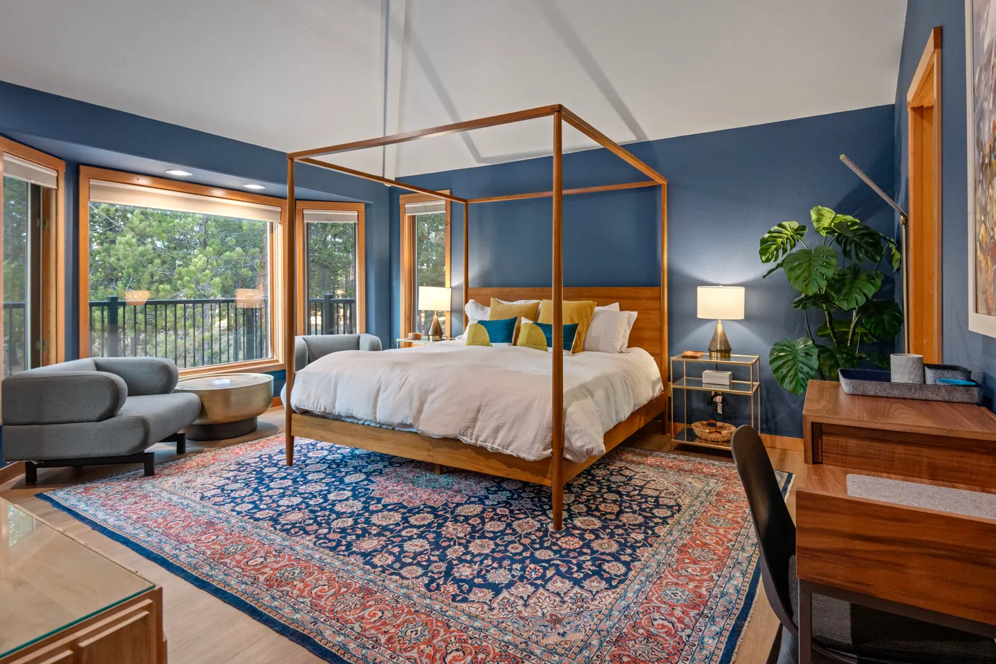 Bedroom featuring a wooden canopy bed on an ornate blue patterned rug, navy blue accent wall, and large windows overlooking trees.