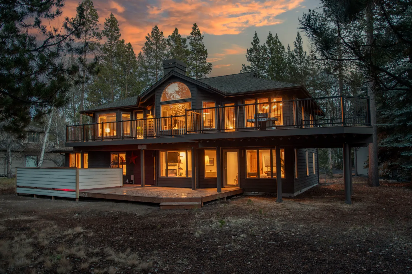 Exterior of a two-story home at twilight with a lower deck, large upper balcony, and glowing interior windows surrounded by pine trees.
