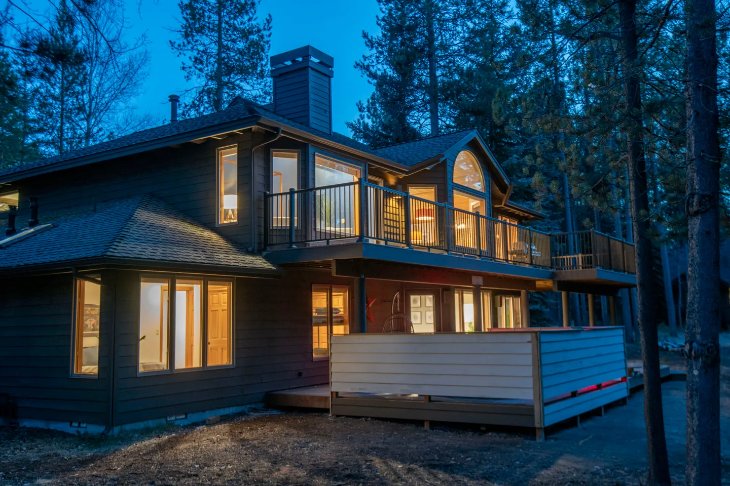 Exterior dusk view of a two-story dark-sided house with an upper deck, lower patio, and glowing interior windows surrounded by pine trees.