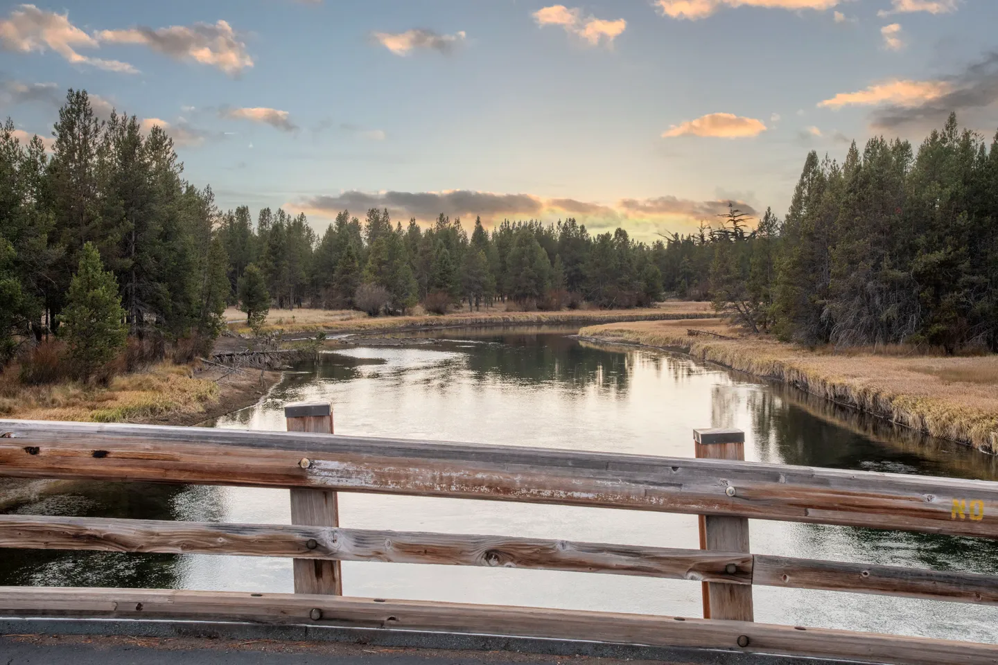 View of a calm river curving through evergreen trees and grassy banks, seen over a wooden bridge railing at dusk.