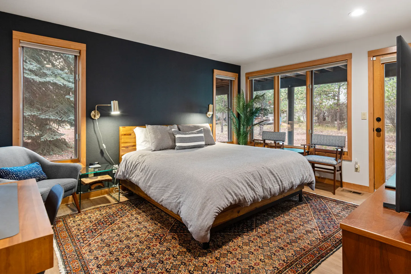 Bedroom featuring a made bed against a dark accent wall, flanked by windows overlooking trees.