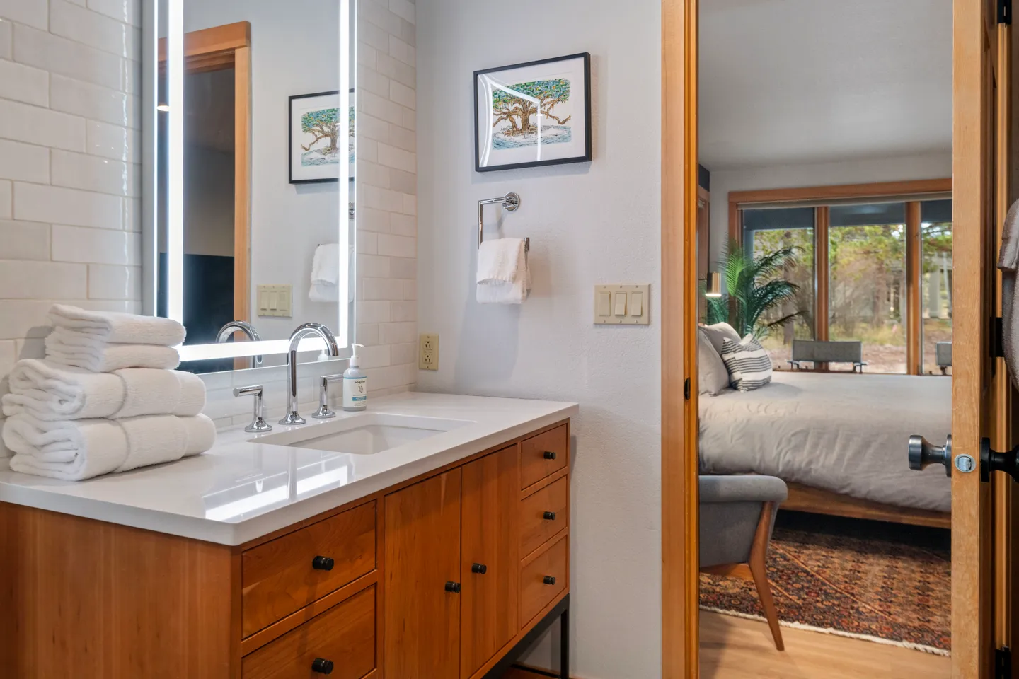 Bathroom vanity with wooden cabinets, white countertop, lighted mirror, and a view into an adjacent bedroom.