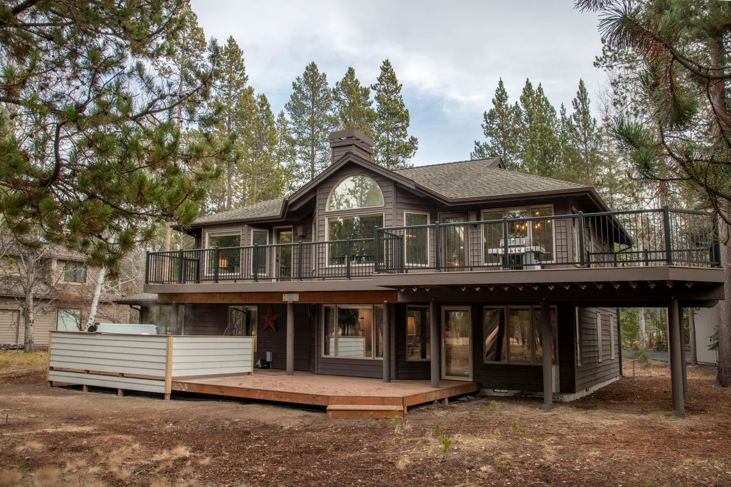 Exterior rear view of a two-story brown wood house with an elevated second-floor deck and a ground-level deck surrounded by pine trees.