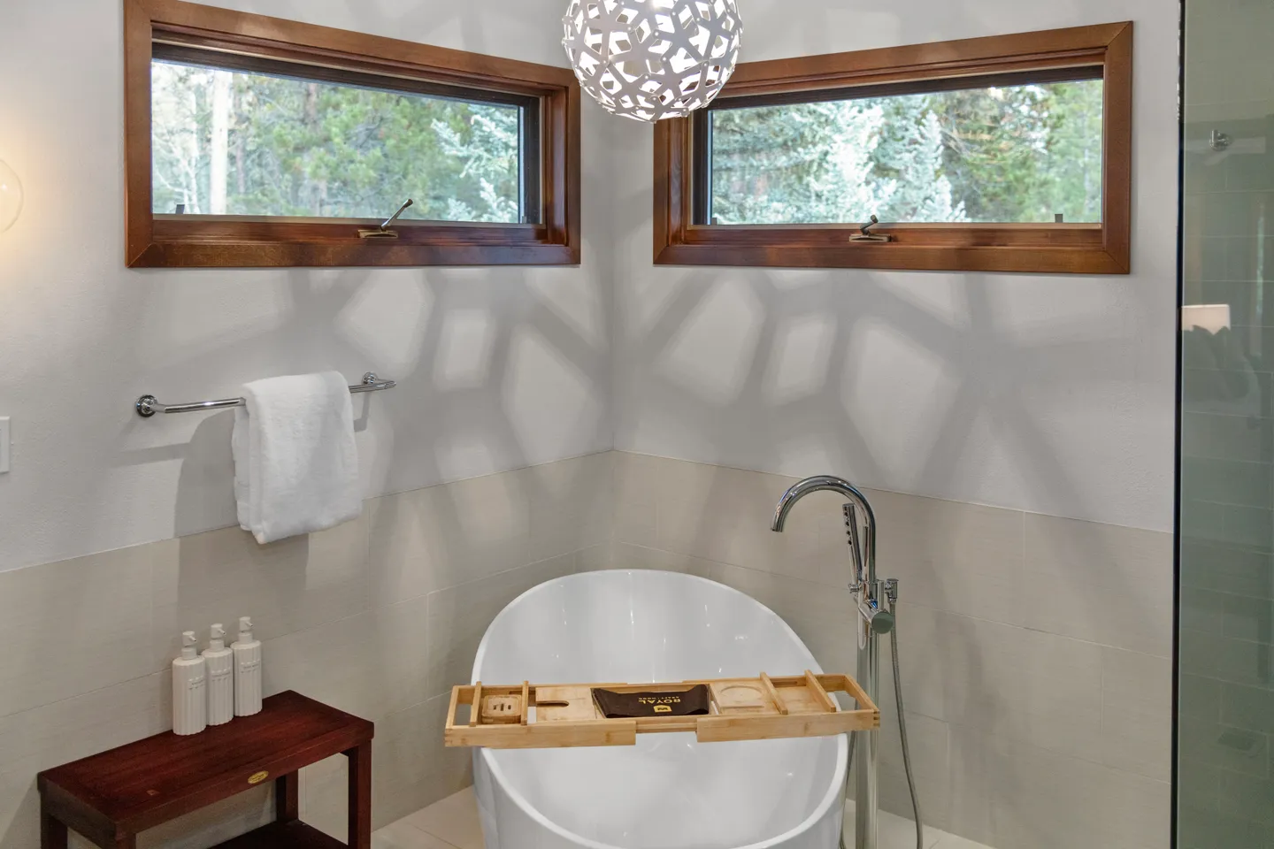 Freestanding soaking tub situated in a bathroom corner beneath awning windows looking out at trees.