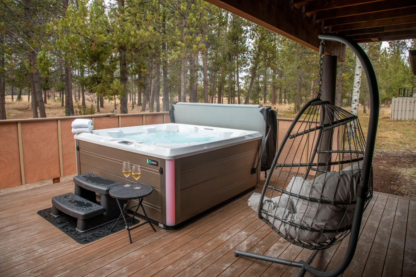 Outdoor wooden deck featuring a steaming hot tub with drinks nearby, next to a hanging egg chair with a forested backdrop.