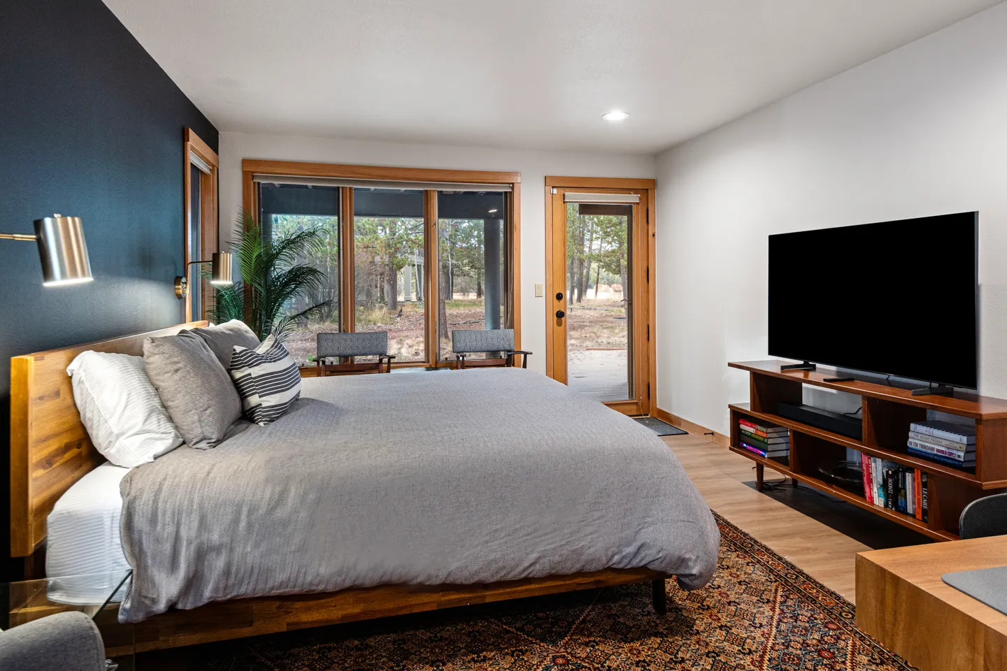 Bedroom featuring a made bed with a dark accent wall, a flat-screen television on a media console, and large windows looking out to trees.