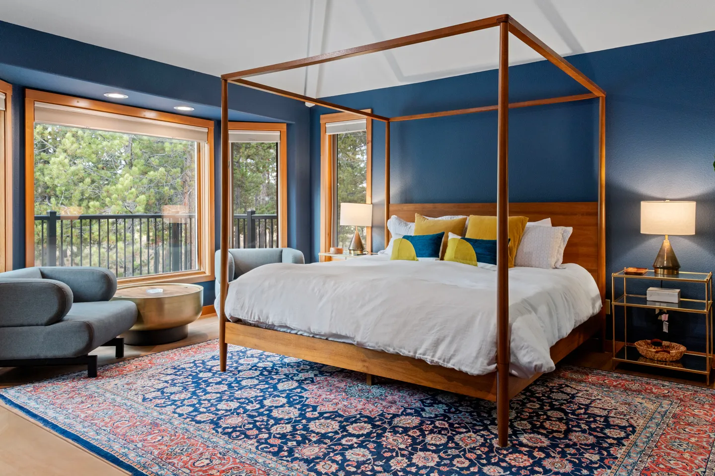 Bedroom with blue walls, a wooden canopy bed, large windows overlooking trees, and an intricate patterned area rug.