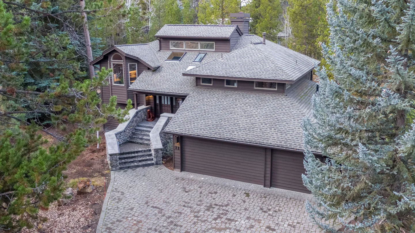 Elevated view of a multi-story wood-sided house surrounded by evergreen trees, featuring a paved driveway and stone front steps.