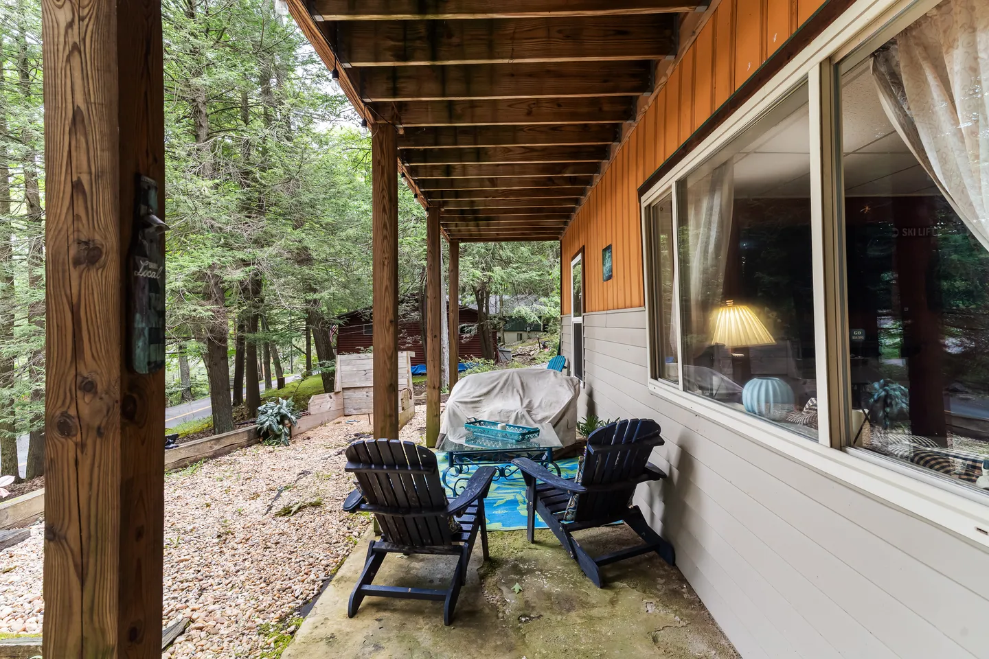 Covered ground-level patio with Adirondack chairs and a small glass-topped table beside a wooded yard.