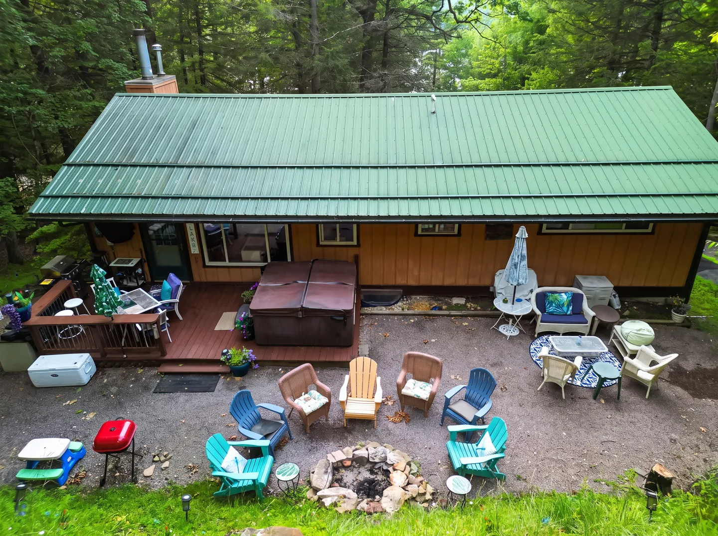 High-angle view of a cabin with a green metal roof, a wooden deck with a covered hot tub, and a gravel yard featuring a stone fire pit surrounded by chairs.
