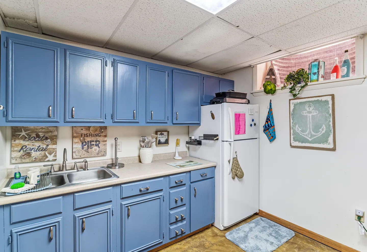 A kitchen with painted blue cabinets, a double sink, light countertops, and a white refrigerator next to a high basement window.