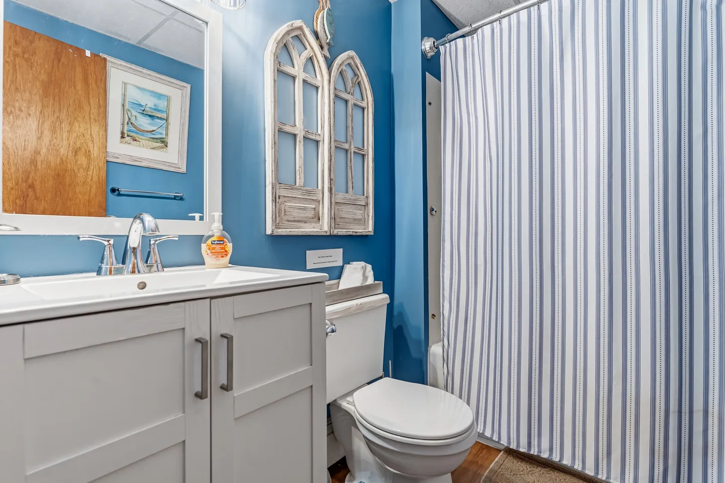 Bathroom with blue walls, a white vanity and sink, a toilet, decorative window-frame wall hangings, and a blue-and-white striped shower curtain.