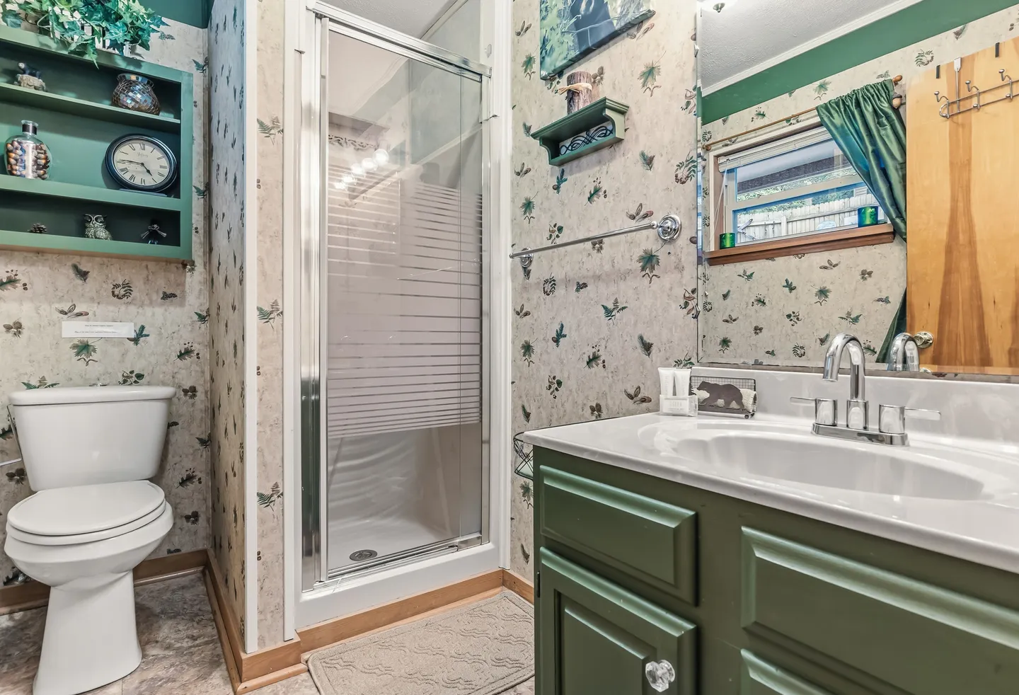 Bathroom with a green vanity, white toilet, patterned wallpaper, and a glass-enclosed shower stall.