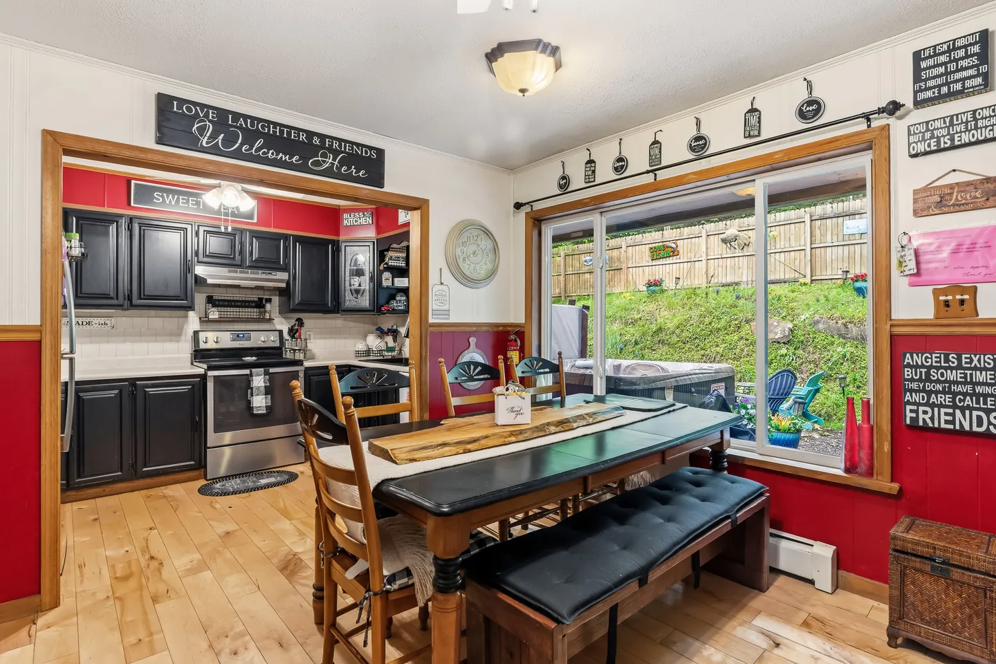 Dining area with a wooden table, bench, and chairs next to large sliding glass doors leading to a patio with a hot tub, opening into a kitchen with black cabinets and stainless steel appliances.