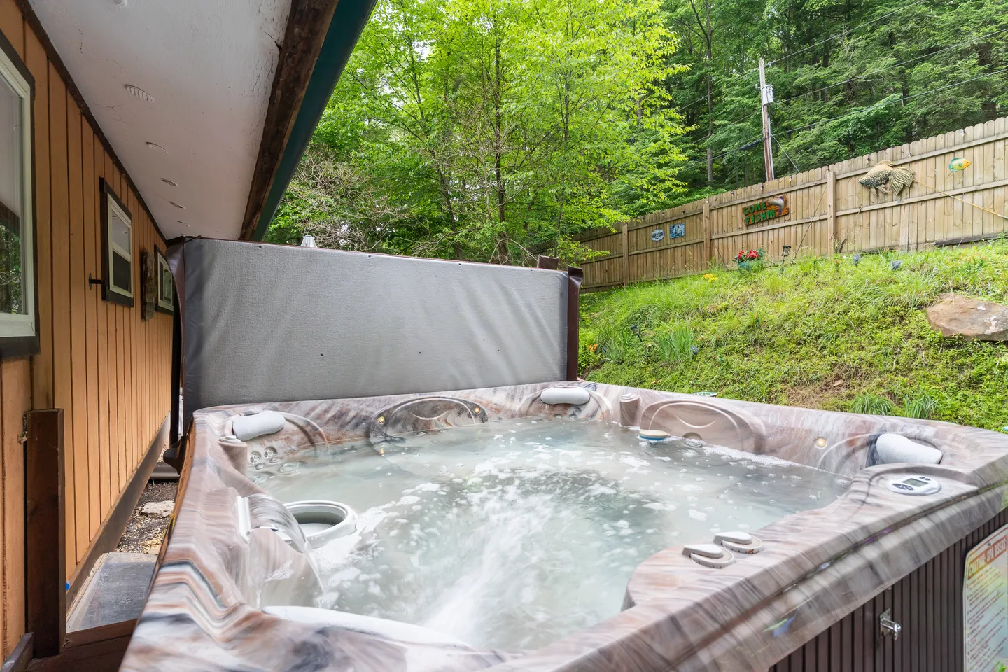 Outdoor bubbling hot tub located next to the house with a privacy cover and a wooden fence along a grassy slope in the background.