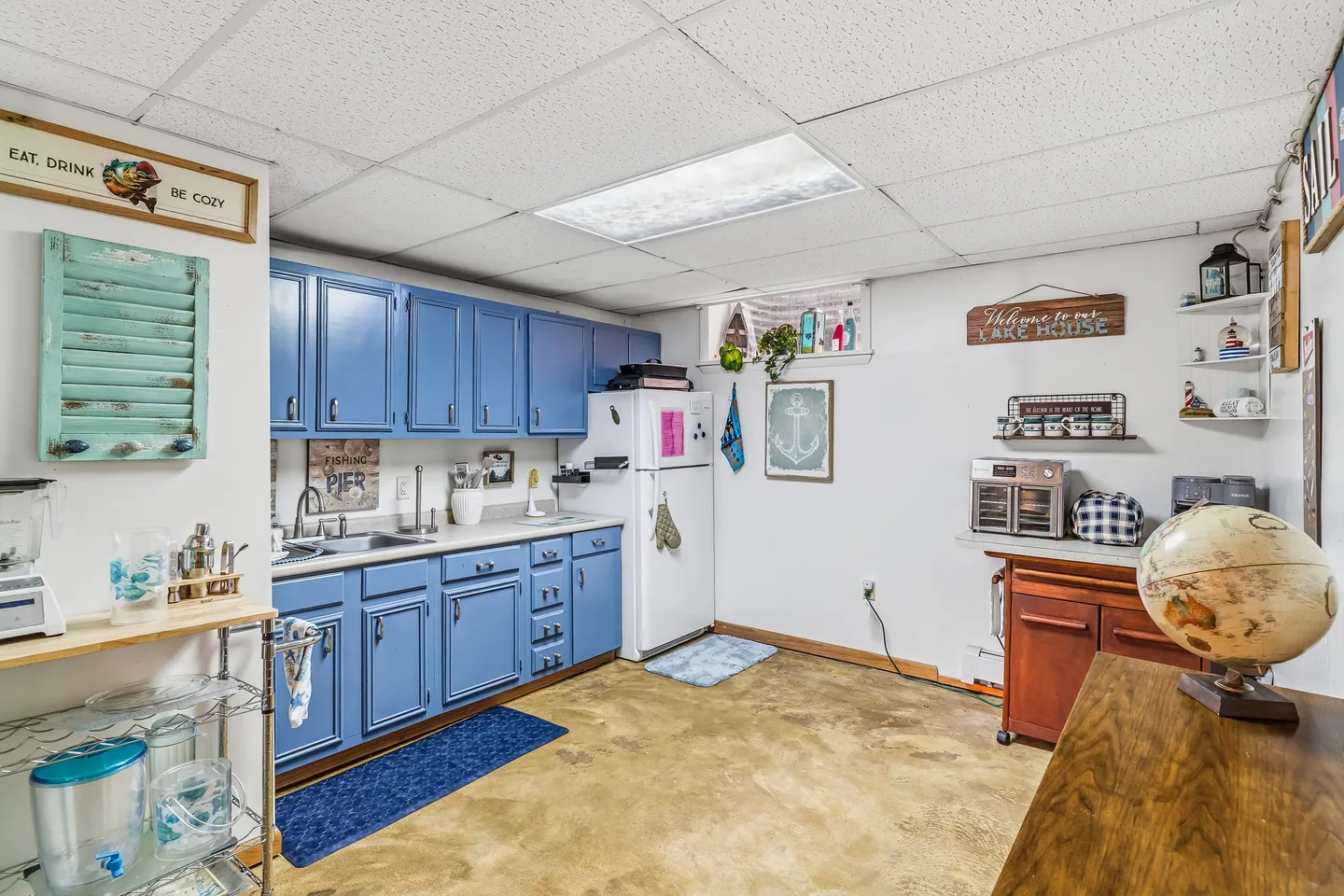 Basement kitchenette with blue cabinetry, a white refrigerator, drop-tile ceiling, and concrete floor.