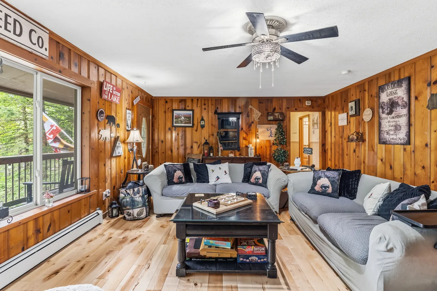 Living room with wood-paneled walls, light wood flooring, two sofas, a dark coffee table, and large window overlooking trees.