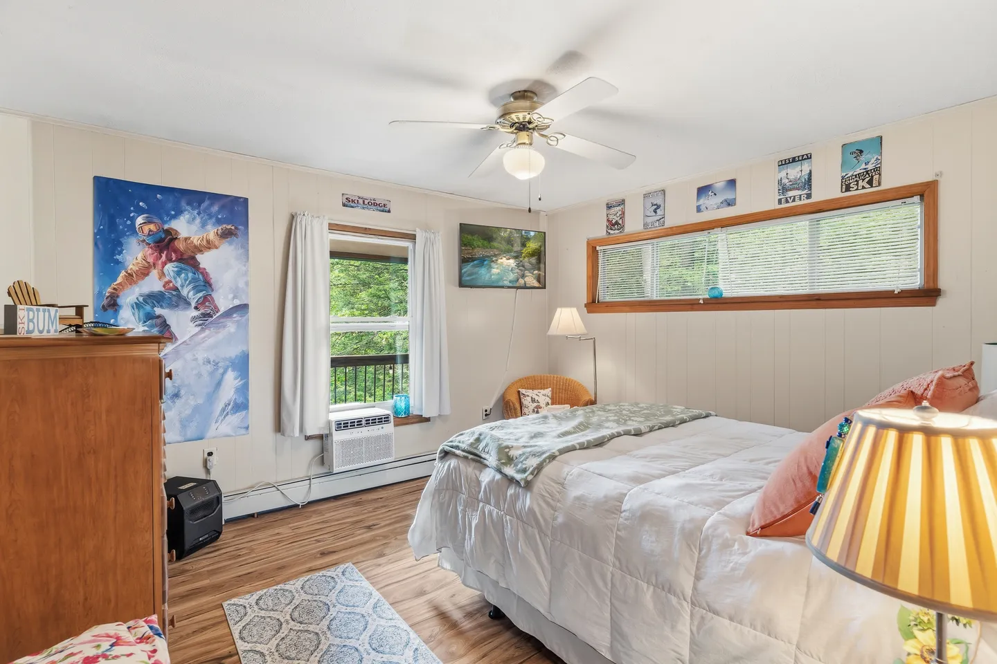 Bedroom with a white-covered bed, wood-style floors, a ceiling fan, windows, and ski-themed wall art.
