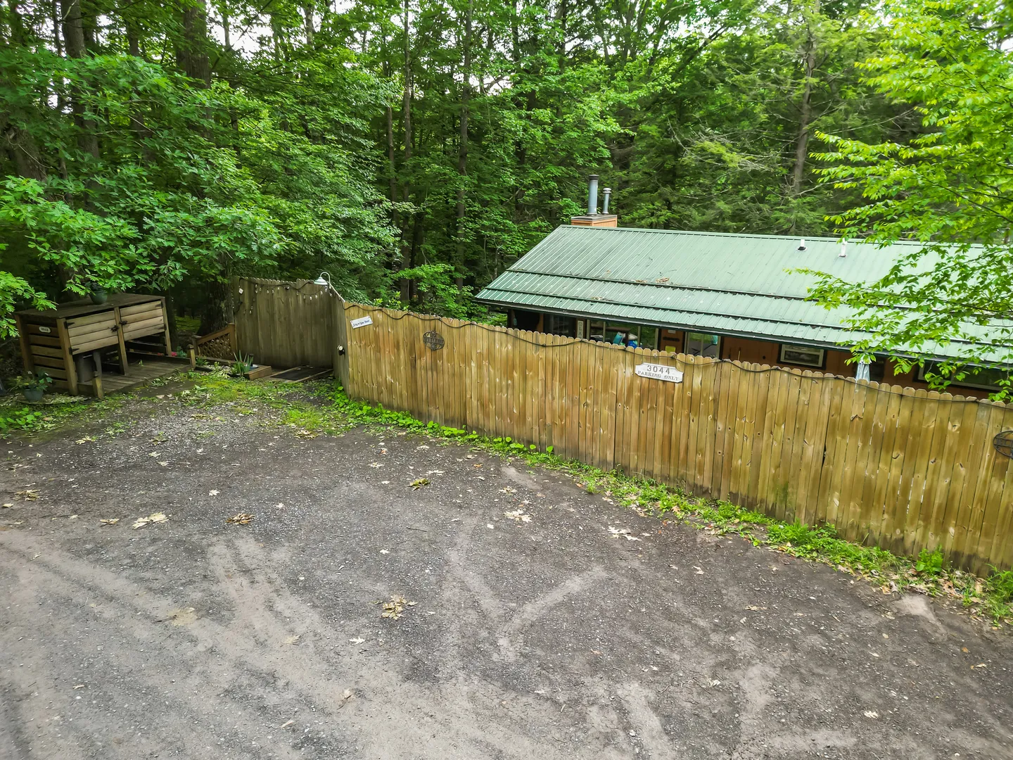 Elevated view of a gravel driveway beside a wooden privacy fence, leading to a cabin nestled in dense woods with a green metal roof.