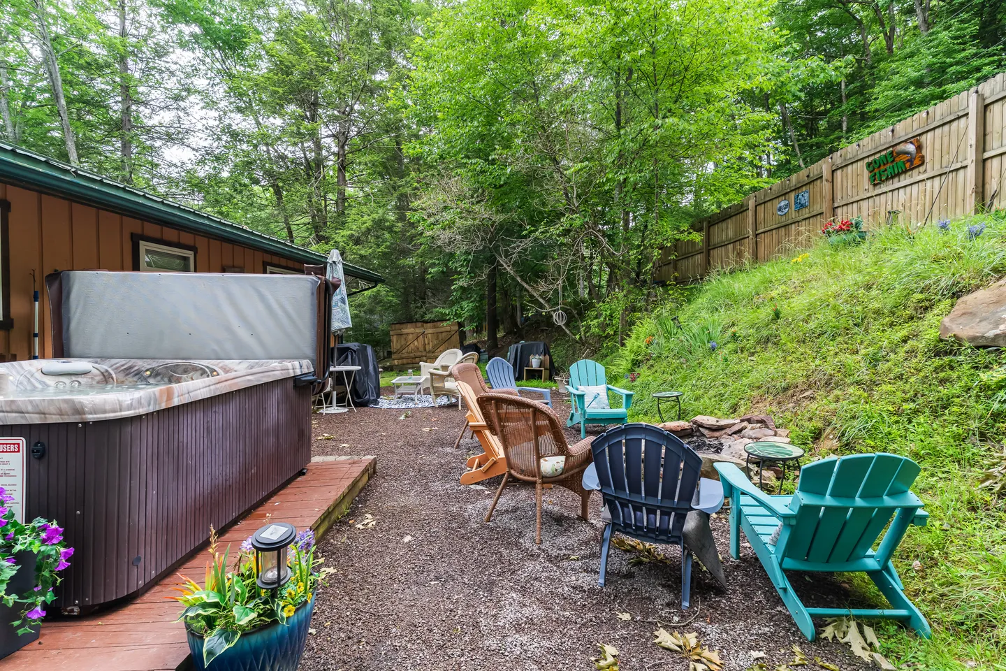 Outdoor backyard area with an open hot tub, gravel patio with Adirondack chairs, and a fire pit near a wooded slope.