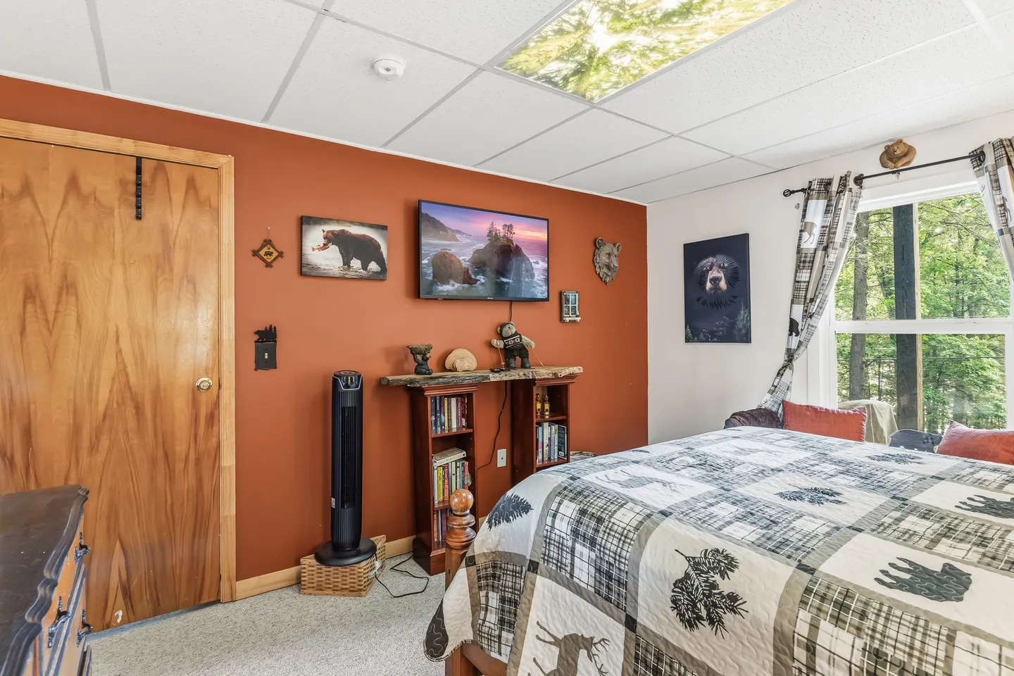 Bedroom featuring a bed with a rustic quilt, a rust-orange accent wall with a wall-mounted TV, and a window viewing wooded greenery.