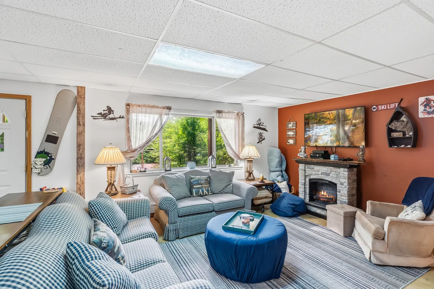 Living room with plaid upholstered seating, a round blue ottoman, a stone-faced fireplace with a wall-mounted TV, and winter sports decor.