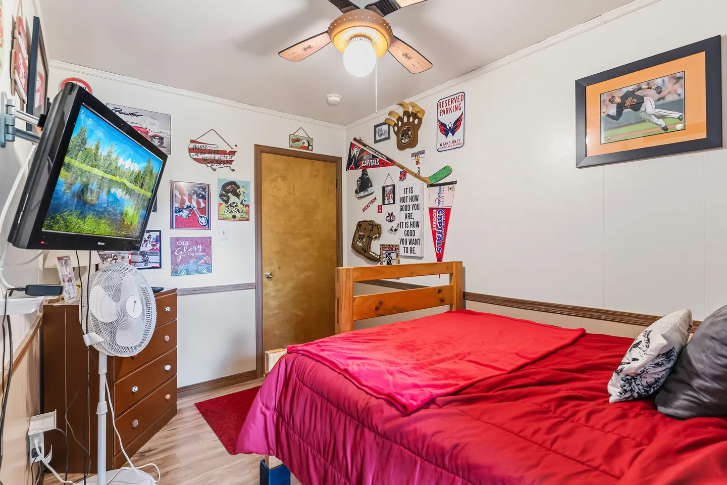 Bedroom with a red comforter on a wooden bed frame, a wall-mounted TV, and sports pennants decorating the wall.