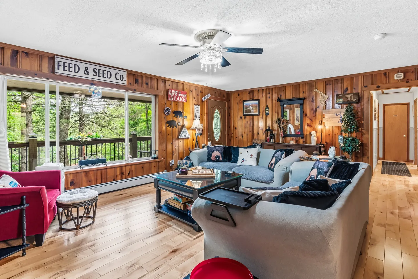 Living room with wood-paneled walls, light wood-look floors, a sectional sofa, coffee table, and large windows looking out to trees.