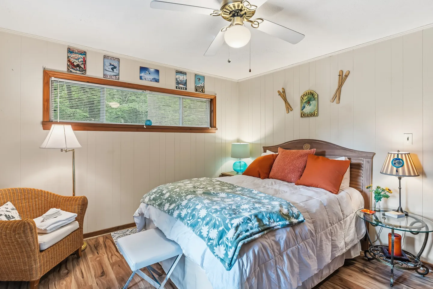 Bedroom with a made bed, wood-paneled walls, ski-themed wall decor, and a horizontal window with blinds.