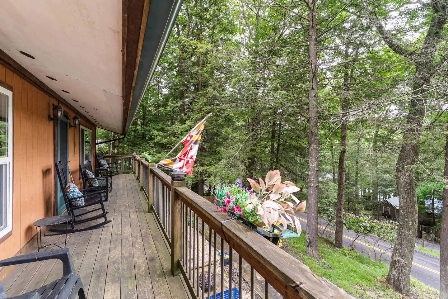 A long covered porch with rocking chairs and railings overlooking tall trees and a roadway.