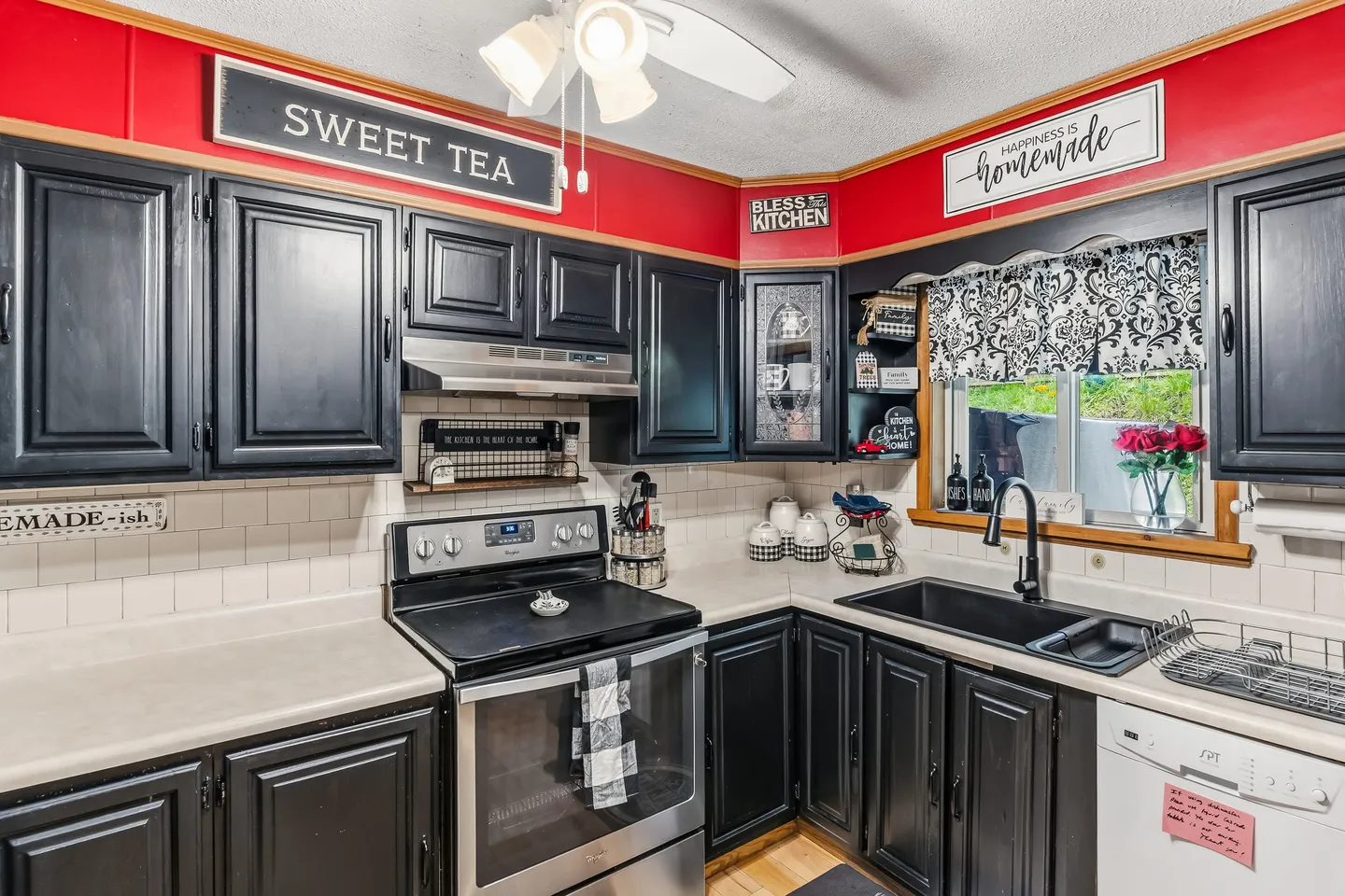 Kitchen with black cabinets, white tiled backsplash, red accent wall trim, stainless steel oven, and a sink under a window.